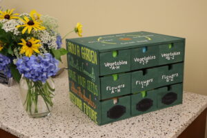 Green box for seeds. There are drawers labeled for flowers, vegetables, and herbs, divided into alphabetical groups. On the side of the box, it says, "Grow a garden. Cultivate a community. Save your seeds. Share the surplus. Feed our future." There is a bouquet of mixed blossoms next to the box.