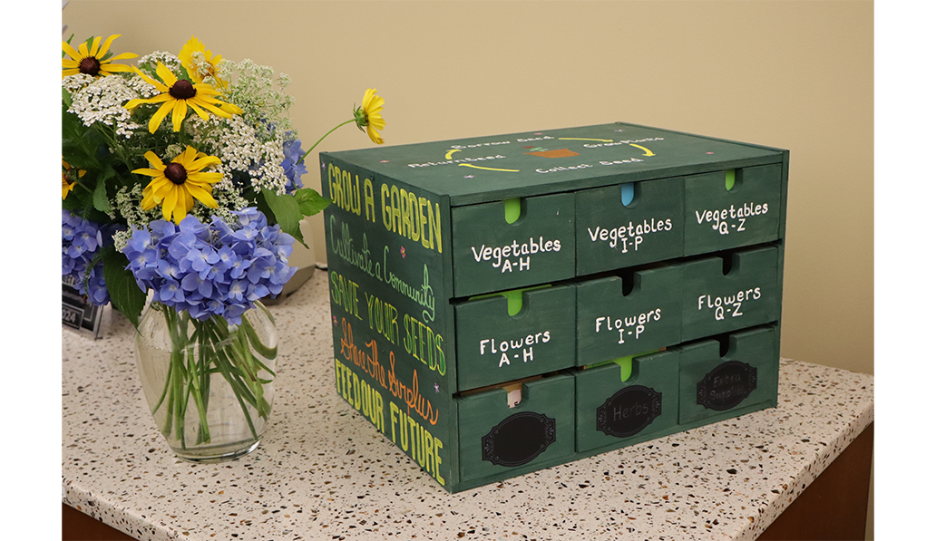 Madison County Library seed library. Green wooden box with drawers that are labeled for seeds. On table with a vase of flowers.