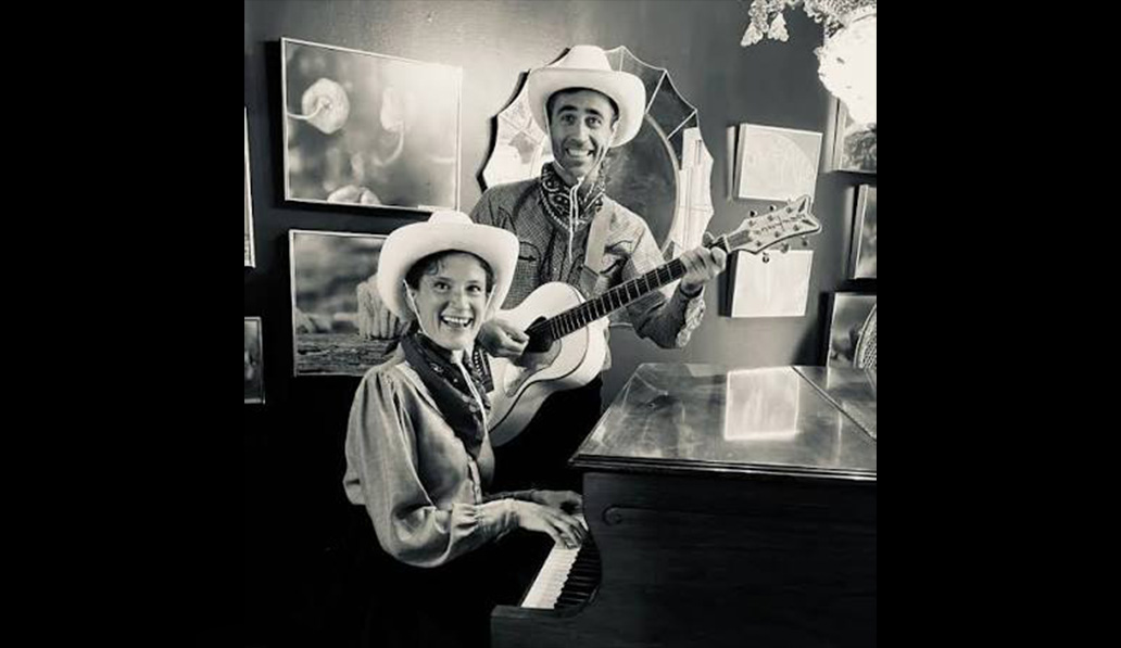 Black and white photo of The PG Show musical duo in western wear. Man playing acoustic guitar and woman playing piano.